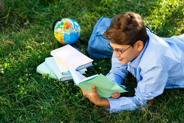 可爱的小男孩 戴着圆眼镜和蓝色衬衣 看书躺在公园的草地上 回到学校概念 — 图库照片
