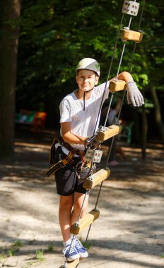 Beyaz t gömlek ve beyaz kask kask ve güvenlik ekipmanları ile macera ip etkinlik parkta sportif, tatlı çocuk. Genç çocuk oynamaya ve sahip eğlenceli aktiviteler açık havada yaptığını. Hobi, aktif yaşam tarzı kavramı
