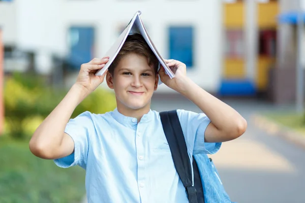 Cute, young boy in blue shirt with backpack stands with workbook on his head in front of his school and smiles. Education, Back to school concept