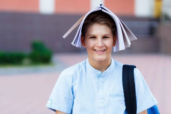 Cute, young boy in blue shirt with backpack stands with workbook on his head in front of his school and smiles. Education, Back to school concept