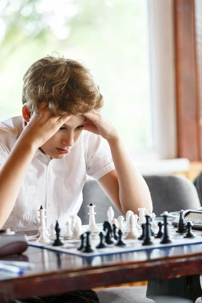 cute, smart, 11 years old boy in white shirt sits in the classroom and plays chess on the chessboard. Training, lesson, hobby, education concept. intellectual game.