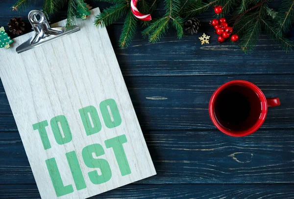 To do list  on wooden pad with red cup of coffee or tea and branches of fir tree, decor on grey wooden table. New Year  Goals list, things to do on Christmas concept. flat lay with copyspace.