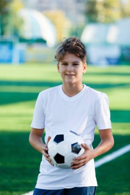 sevimli genç çocuk beyaz mavi spor Stadyumu alan yeşil çimenlerin üzerinde klasik siyah ve beyaz futbol topu tutuyor. Eğitim, maç, hobi kavramı. kopya alanı ile