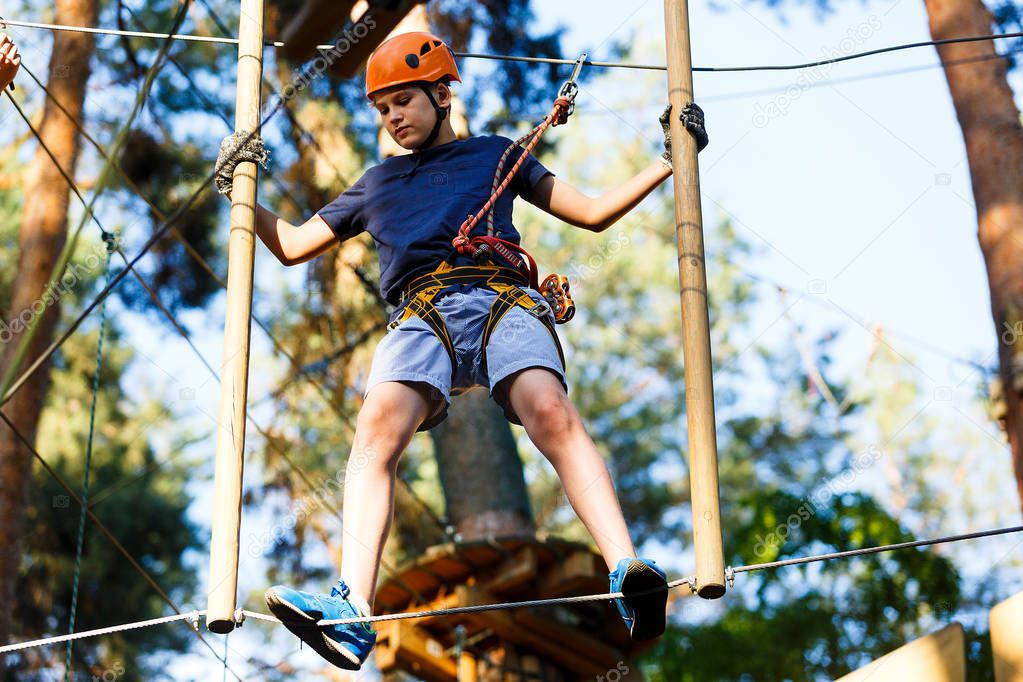 Child in forest adventure park. Kid in orange helmet  and blue t shirt climbs on high rope trail. Agility skills and climbing outdoor amusement center for children. young boy plays outdoors.