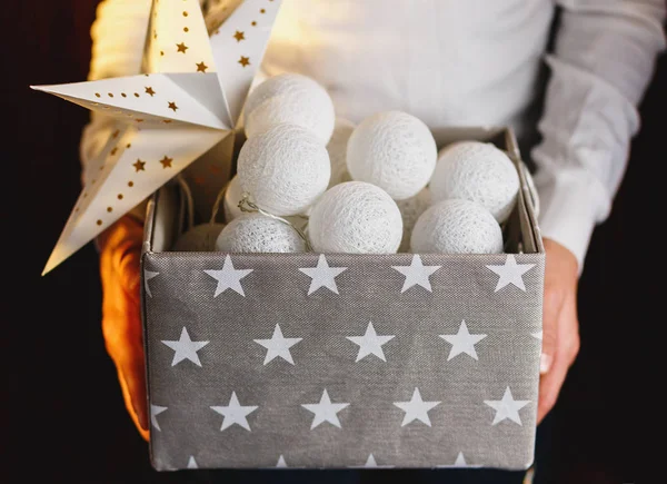 White cotton ball light garland in a grey basket on boys hands with stars sparkling at home. close up, bright lights, festive atmosphere. home decoration. New year and Christmas celebration concept