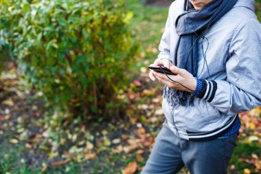 gri eşarp ile gri ceketli genç çocuk tutan ve sonbahar arka plan üzerinde kulaklık dışında ile smartphone kullanır. Teknoloji, sosyal medya smartphone içinde 