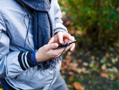 gri eşarp ile gri ceketli genç çocuk tutan ve sonbahar arka plan üzerinde kulaklık dışında ile smartphone kullanır. Teknoloji, sosyal medya smartphone içinde 