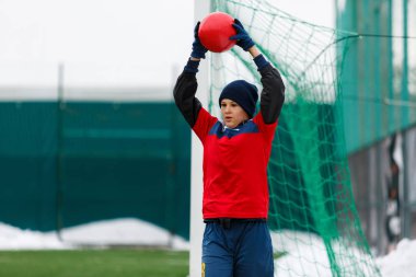 Genç aktif yakışıklı kırmızı ve mavi spor giyim kış alan arka plan üzerinde kar ile gelen bir futbol topu atmak. kış aktiviteleri, futbol oyun, eğitim kavramı