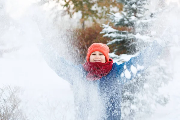 cute young boy in orange hat red scarf and blue jacket plays with snow, has fun, smiles. Teenager rises his hands up in winter park. Active lifestyle, winter activity, outdoor winter games, snowballs