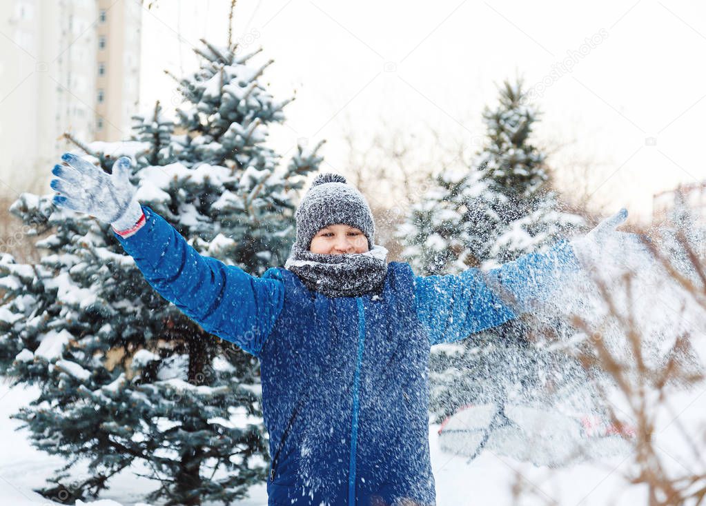cute young boy in orange hat red scarf and blue jacket plays with snow, has fun, smiles. Teenager rises his hands up in winter park. Active lifestyle, winter activity, outdoor winter games, snowballs