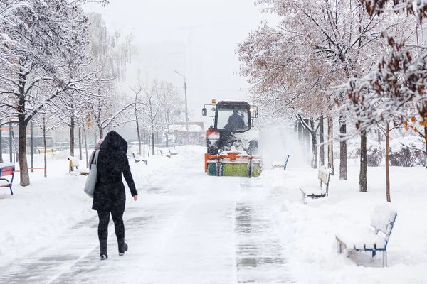 Snow clearing. Tractor clears road, way after heavy snowfall. Tractor cleaning the road from the snow. Woman walking along the alley of a snowy park.