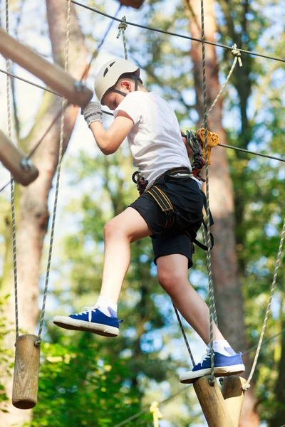 Child in forest adventure park. Kid in white helmet and white t shirt climbs on high rope trail. Agility skills and climbing outdoor amusement center for children. young boy plays outdoors