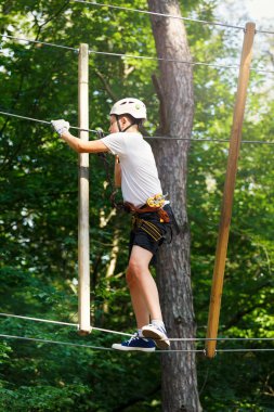 Orman macera parkı çocuk. Çocuk turuncu kask ve mavi t gömlek tırmanıyor yüksek ip izinde. Çeviklik beceri ve açık hava eğlence merkezi çocuklar için tırmanma. Genç çocuk açık havada çalış