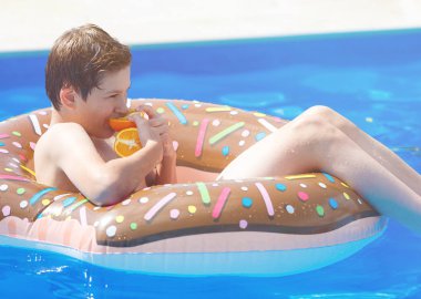 Cute child boy on funny inflatable donut float ring in swimming pool with oranges. Teenager learning to swim, has fun in outdoor pool at resort. Water toys for kids. Healthy sport activity, vacation 