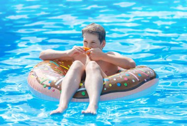Cute child boy on funny inflatable donut float ring in swimming pool with oranges. Teenager learning to swim, has fun in outdoor pool at resort. Water toys for kids. Healthy sport activity, vacation 