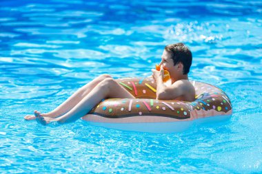 Cute child boy on funny inflatable donut float ring in swimming pool with oranges. Teenager learning to swim, has fun in outdoor pool at resort. Water toys for kids. Healthy sport activity, vacation 