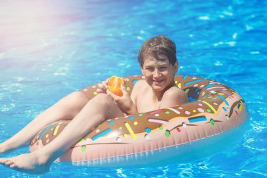 Cute child boy on funny inflatable donut float ring in swimming pool with oranges. Teenager learning to swim, has fun in outdoor pool at resort. Water toys for kids. Healthy sport activity, vacation 