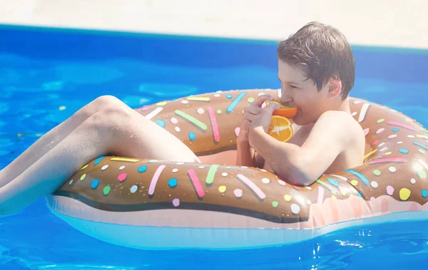 Cute child boy on funny inflatable donut float ring in swimming pool with oranges. Teenager learning to swim, has fun in outdoor pool at resort. Water toys for kids. Healthy sport activity, vacation 