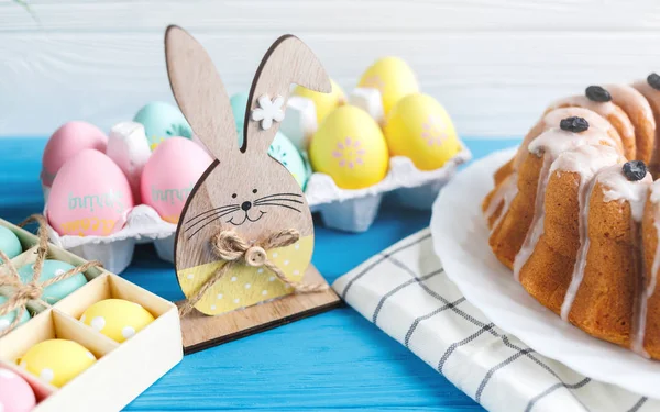 Happy Easter! Handmade cake on towel, eggs, wooden bunny rabit on blue wooden background. Decoration for Easter holiday