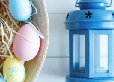 Mutlu Paskalyalar! Elle boyanmış renkli yumurtalar ve beyaz ahşap masa üzerinde mavi fenerle bir kase. Kapatın. Paskalya için dekorasyon, şenlik arka planı.