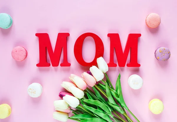 Word Mum from red letters and tender tulips, macaroons on pink background. Mothers day decoration concept. Top view, flat lay with copy space