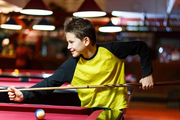 Cute boy in yellow t shirt plays billiard or pool in club. Young Kid learns to play snooker. Boy with billiard cue strikes the ball on table. Active Leisure, sport, hobby concept