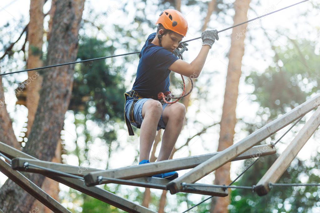 Sporty, young, cute boy in white t shirt spends his time in adventure rope park in helmet and safe equipment in the park in the summer. Active lifestyle concept
