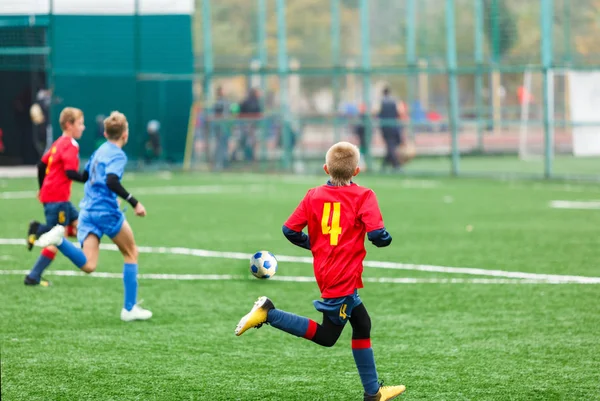 Young footballers dribble and kick football ball in game. Boys in red  blue sportswear running on soccer field. Training, active lifestyle, sport, children activity concept