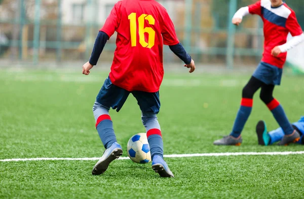 Young footballers dribble and kick football ball in game. Boys in red  blue sportswear running on soccer field. Training, active lifestyle, sport, children activity concept