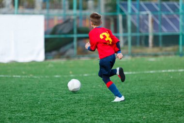 Kırmızı ve mavi spor giyen çocuklar sahada futbol oynar, top top akıtır. Yeşil çimüzerinde topu ile genç futbolcular. Eğitim, futbol, çocuklar için aktif yaşam tarzı 