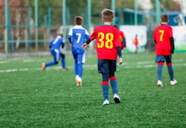 Kırmızı ve mavi spor giyen çocuklar sahada futbol oynar, top top akıtır. Yeşil çimüzerinde topu ile genç futbolcular. Eğitim, futbol, çocuklar için aktif yaşam tarzı 