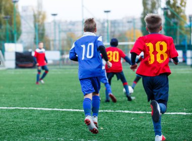 Kırmızı ve mavi spor giyen çocuklar sahada futbol oynar, top top akıtır. Yeşil çimüzerinde topu ile genç futbolcular. Eğitim, futbol, çocuklar için aktif yaşam tarzı 