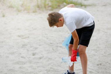 Eldivenli beyaz tişörtlü çocuk sahilde ki mavi pakette çöp ve plastik şişeleri topluyor. Genç gönüllü. Çevre koruma, çevre kavramını koruma. 
