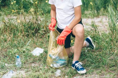 Eldivenli beyaz tişörtlü çocuk sahilde ki mavi pakette çöp ve plastik şişeleri topluyor. Genç gönüllü. Çevre koruma, çevre kavramını koruma. 
