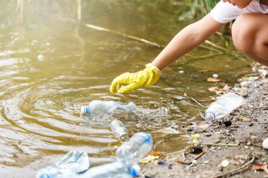 Eldivenli beyaz tişörtlü çocuk sahilde ki mavi pakette çöp ve plastik şişeleri topluyor. Genç gönüllü. Çevre koruma, çevre kavramını koruma. 