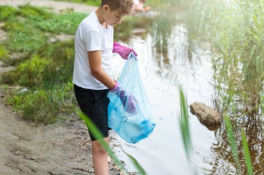 Eldivenli beyaz tişörtlü çocuk sahilde ki mavi pakette çöp ve plastik şişeleri topluyor. Genç gönüllü. Çevre koruma, çevre kavramını koruma. 