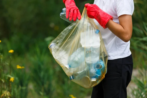 Eldivenli beyaz tişörtlü çocuk sahilde ki mavi pakette çöp ve plastik şişeleri topluyor. Genç gönüllü. Çevre koruma, çevre kavramını korumak. Eldivenle ellerinizi kapatın