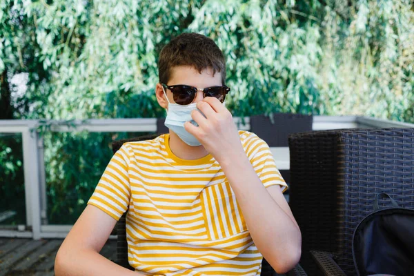 stock image Cute teenager in yellow t shirt and mask on his face in city cafe. Boy wearing protective mask while waiting his order at open terrace of restaurant. Stay safe outdoor.