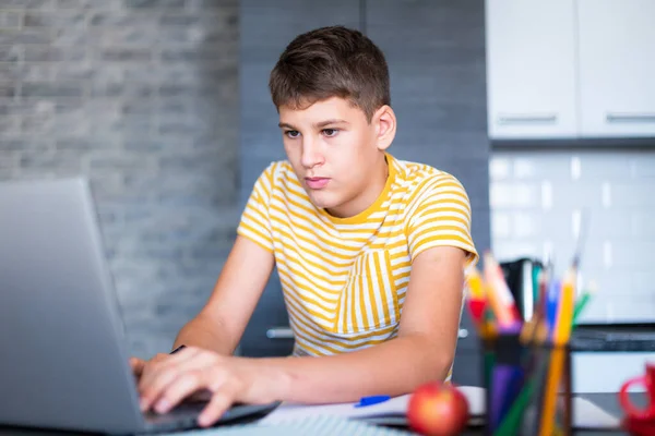 Cute young teenager in white shirt sitting behind desk in kitchen next to laptop and study. Serious boy in earphones makes homework, listening lesson. Home, distance education, self study by kids.