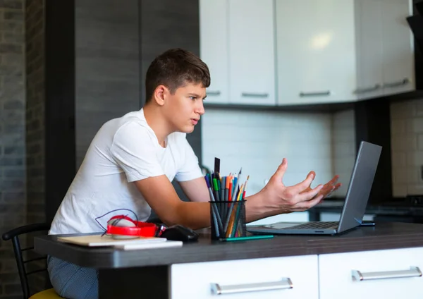 Cute young teenager in white shirt sitting behind desk in kitchen next to laptop and study. Serious boy makes homework, listening lesson. Home, distance education, self study by kids.