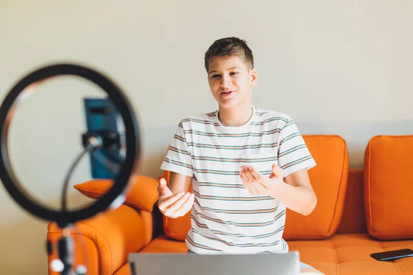 Young teenager blogger recording video at home. Cheerful cute boy pointing with hands into a smartphone. Boy with camera recording video on the orange couch. Social media concept.