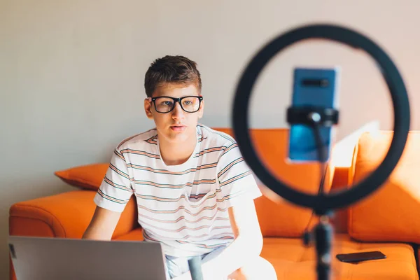 Young teenager blogger recording video at home. Cheerful cute boy pointing with hands into a smartphone. Boy with camera recording video on the orange couch. Social media concept.