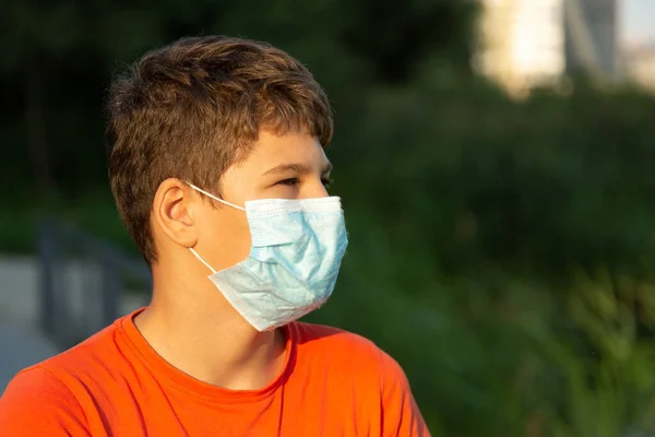 彼の顔に保護マスクでかわいい少年を閉じます コロナウイルス 感染から保護するために医療マスクを身に着けている白いTシャツのティーンエイジャー 街中の隔離 — ストック写真