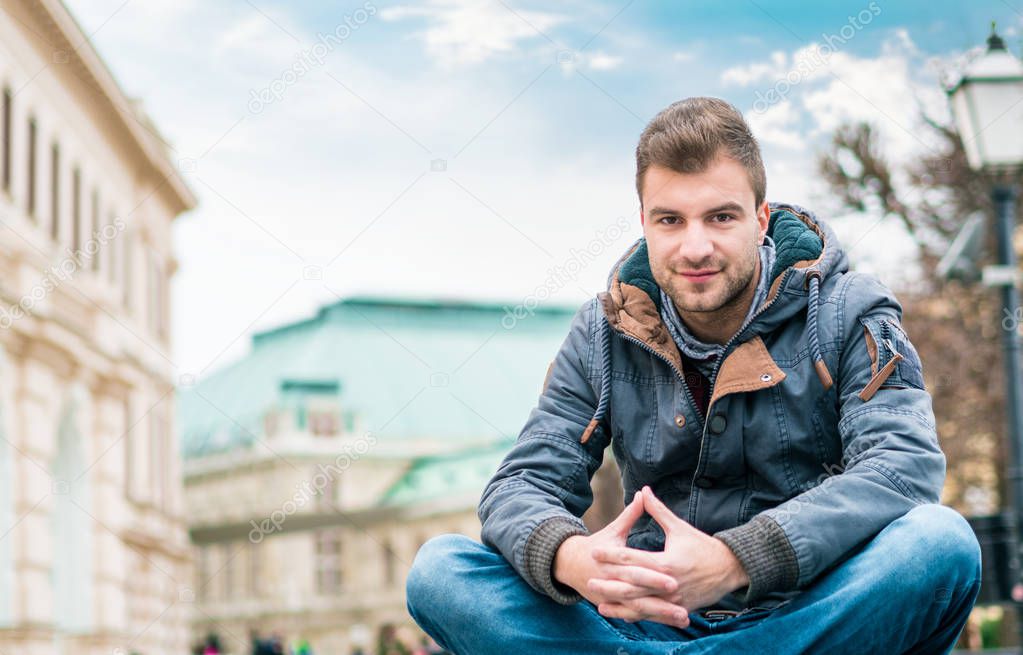 Young handsome man sitting. Gorgeous guy looking at camera in front of old city