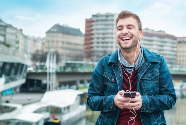 快乐的家伙在他的手机上听音乐 微笑的年轻人在 Earpones 在模糊的城市前面 — 图库照片