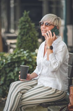 40 'lı yaşlarda orta yaşlı sarışın bir kadın cep telefonuyla şehirde bankta konuşuyor ve dinleniyor. Yetişkin kadınların akıllı telefondaki konuşmalarının profili veya yan görünümü.