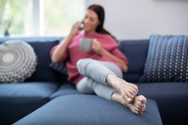 Mujer joven que relaja en el sofá con la bebida caliente que habla en móvil Ph — Foto de Stock