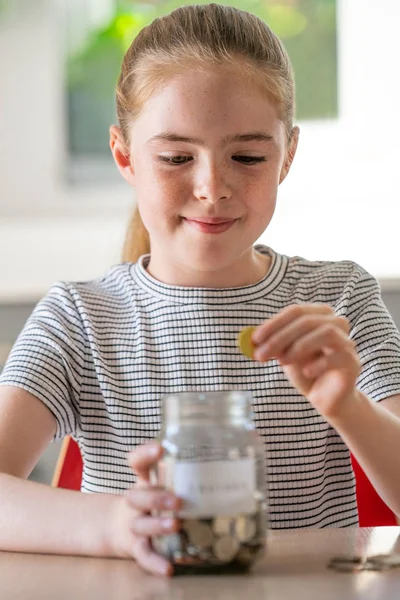 Chica poniendo monedas en tarro de vidrio etiquetado ahorros en casa —  Fotos de Stock