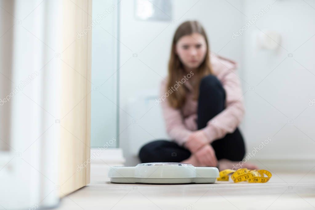 Unhappy Teenage Girl Sitting In Bathroom Looking At Scales And T