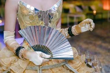 Işıldayan altın elbisesi içinde bir dansçı zarif bir şekilde gümüş yelpaze tutuyor ve akşam kutlamalarına katılıyor..